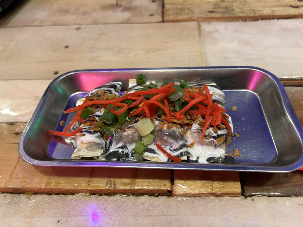 Quatre boulettes longues et étroites disposées les unes à côté des autres sur un plateau en métal, recouvertes de sauce blanche et noire et garnies de gingembre mariné râpé rouge vif.