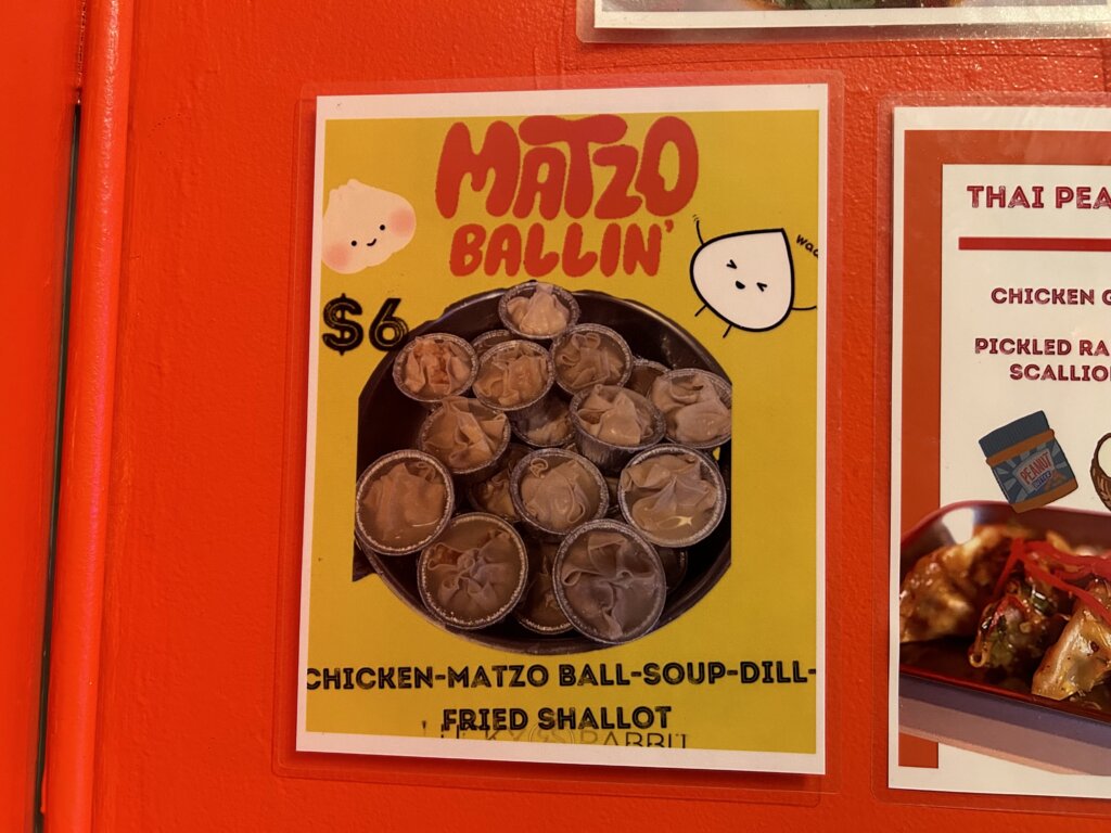 Une affiche avec les mots "Matzo Ballin'" en haut, au-dessus d'une pile de petits verres à shot en aluminium remplis de raviolis recouverts de bouillon et garnis d'échalotes frites et d'aneth.  En bas, les mots "poulet-boule de matzo-souop-échalote frite à l'aneth."