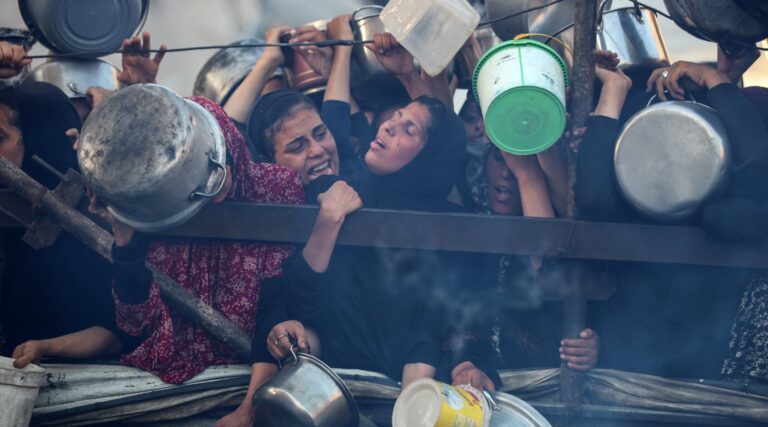 Palestinians gathered outside of an aid site carrying pots, one woman looking up in distress.